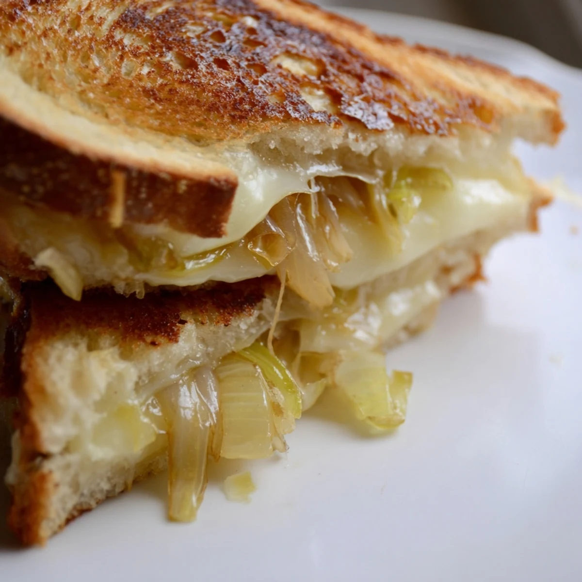 Golden-brown Havarti & Caramelized Leek Grilled Cheese slices are stacked beside a bowl of creamy tomato soup on a rustic table.  